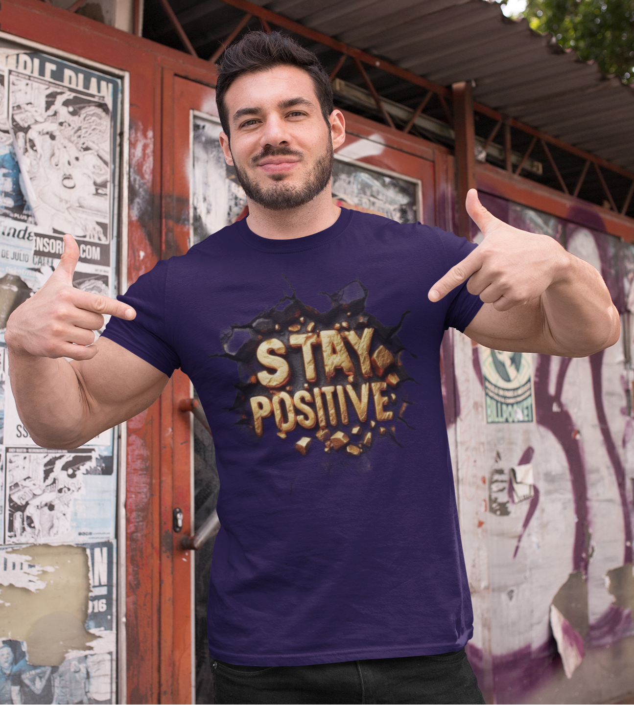 Man wearing a purple t-shirt with 'Stay Positive' text, giving a thumbs-up gesture in front of a graffiti-covered wall.